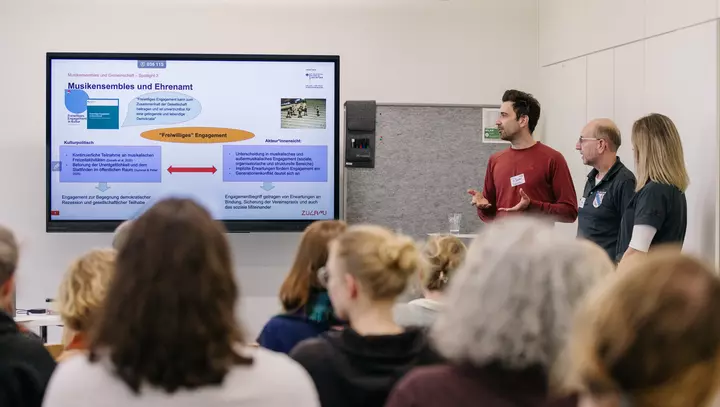 3 Personen präsentieren vor Zuschauenden neben einem Bildschirm. 