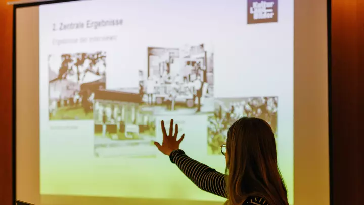 Eine Frau steht vor einer Leinwand, auf die eine Präsentationsfolie projiziert ist.