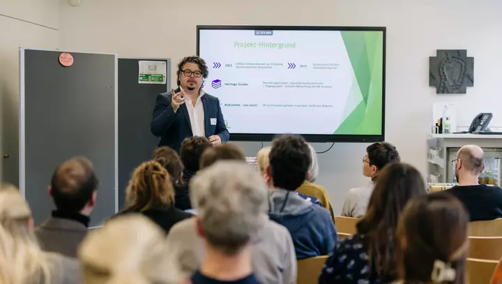 Ein Mann präsentiert vor Zuschauenden neben einem Bildschirm, auf dem eine Präsentationsfolie zu sehen ist.