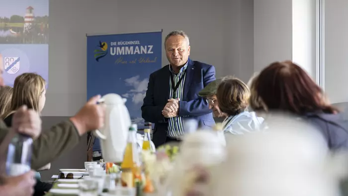 Ein Mann steht am Ende eines Tisches, an dem mehrere Personen sitzen. 