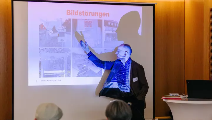 Ein Mann steht vor einer Leinwand, auf die eine Präsentationsfolie projiziert ist. 