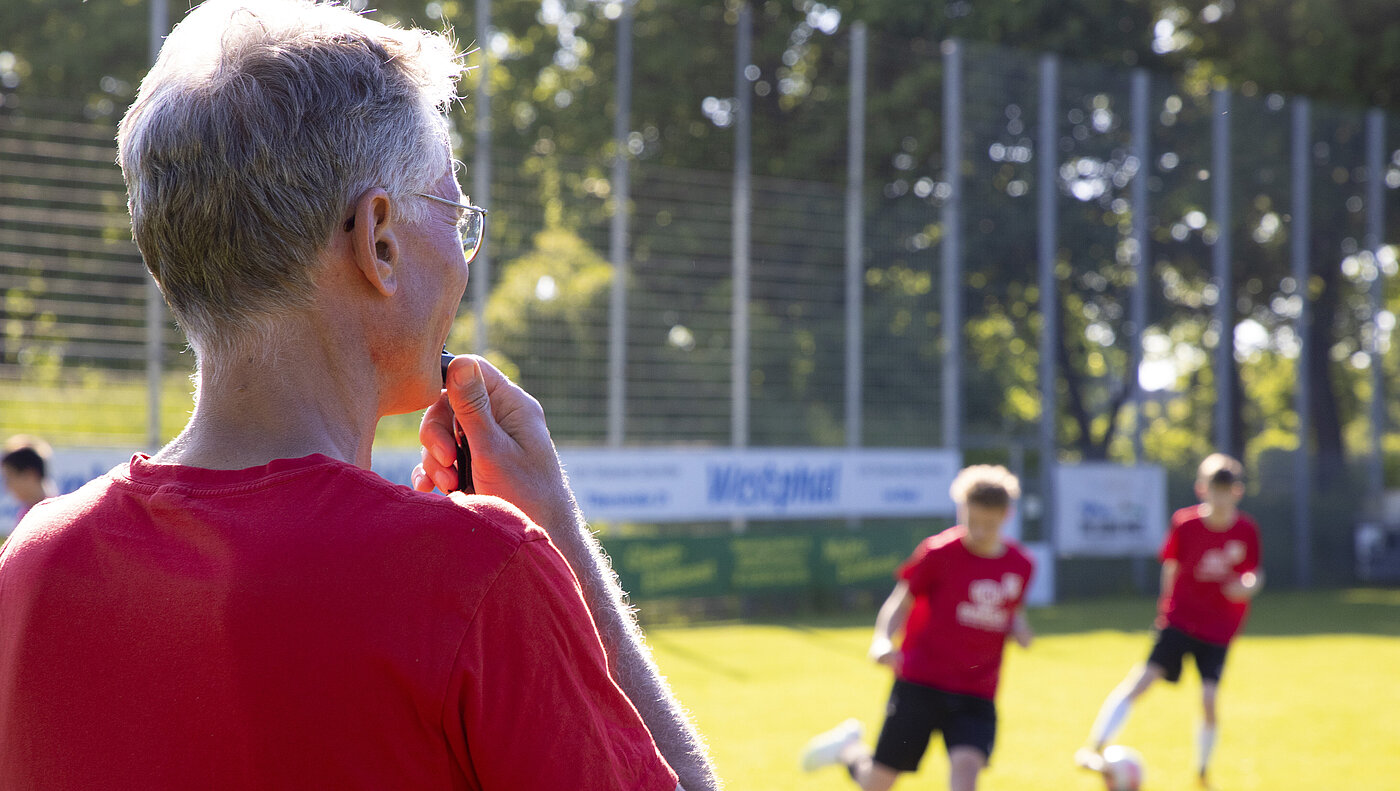 Ein Trainer mit Trillerpfeife sowie zwei fußballspielende Kinder auf einem Fußballfeld