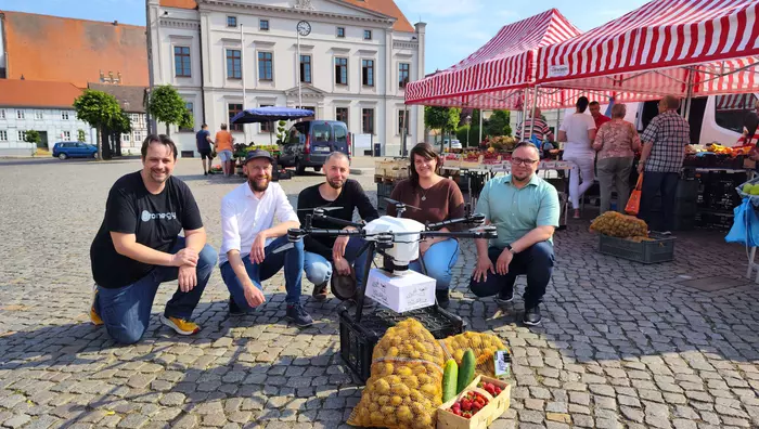 Das fünfköpfige Projektteam von Stadt-Land-Drohne kniet auf dem Wusterhausener Marktplatz hinter einer Lieferdrohne, im Hintergrund ist das Rathaus zu sehen.