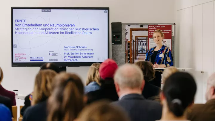 Eine Frau präsentiert vor Zuschauenden neben einem Bildschirm, auf dem eine Präsentationsfolie zu sehen ist. 