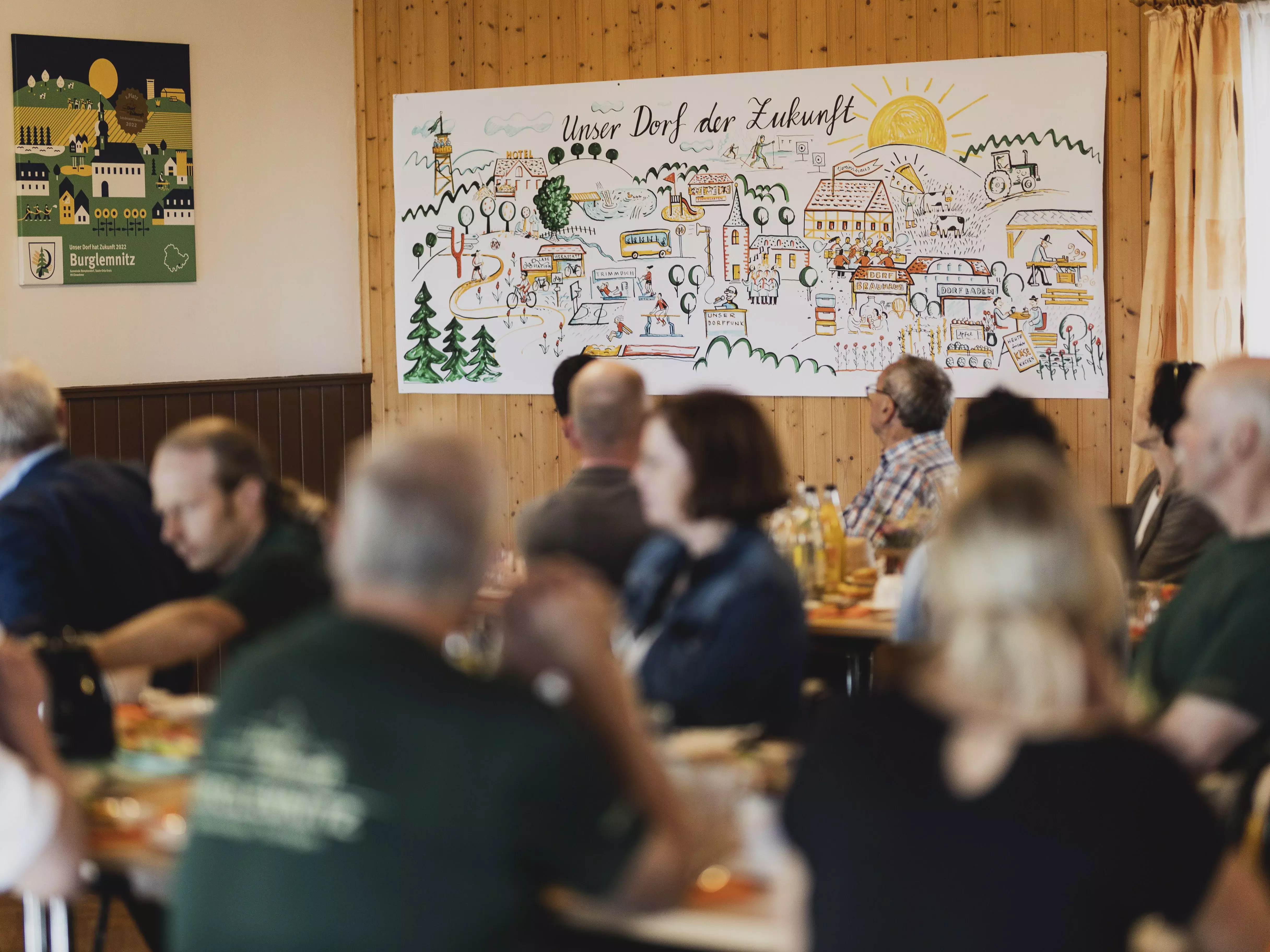 Im Vordergrund sitzen Menschen an Tischen, im Hintergrund ist ein Plakat mit der Überschrift "Unser Dorf der Zukunft" aufgehängt