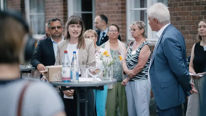 Mehrere Person stehen verteilt an runden Stehtischen. Im Vordergrund ist Lara Grubert, Projektkoordinatorin von "Wir in Serwest" im Gespräch mit Bundesminister Alois Rainer zu sehen.