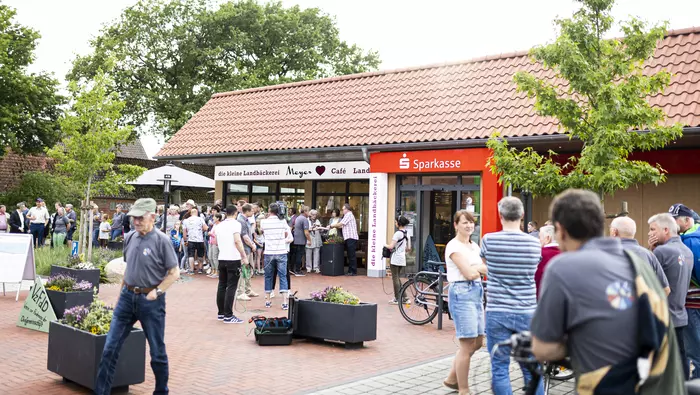 Menschen stehen in Grüppchen auf einem Dorfplatz mit Bäckerei und Sparkasse