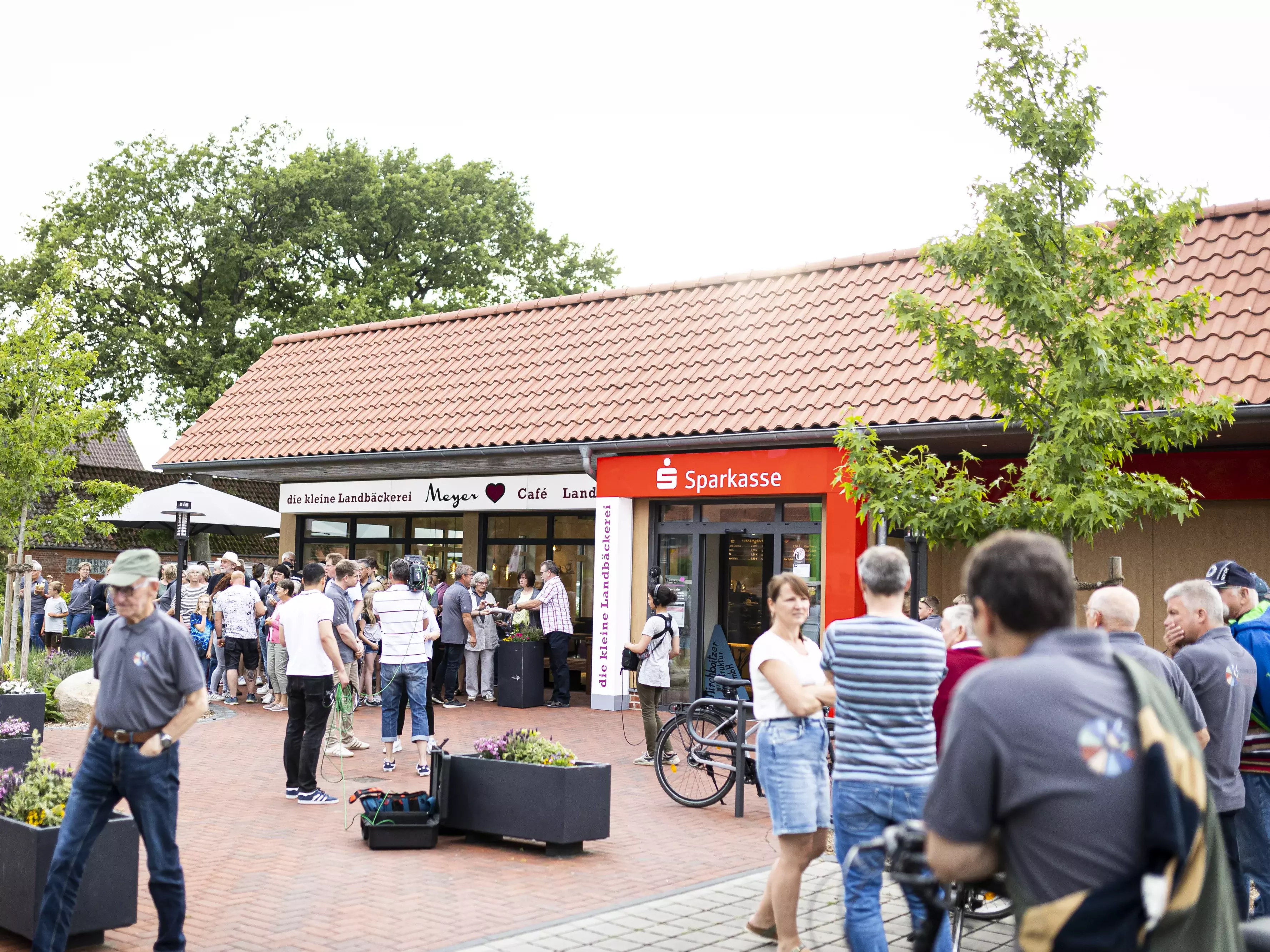 Menschen stehen in Grüppchen auf einem Dorfplatz mit Bäckerei und Sparkasse