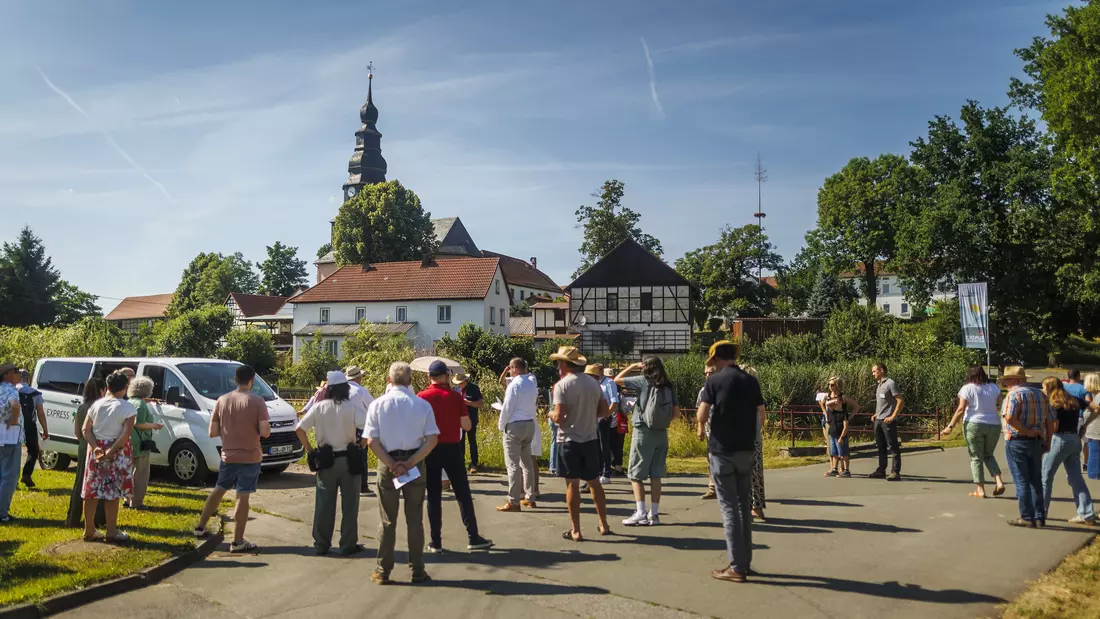 Eine Menschenansammlung steht im Sonnenschein vor einer dörflichen Kulisse und hört einem Redner zu.