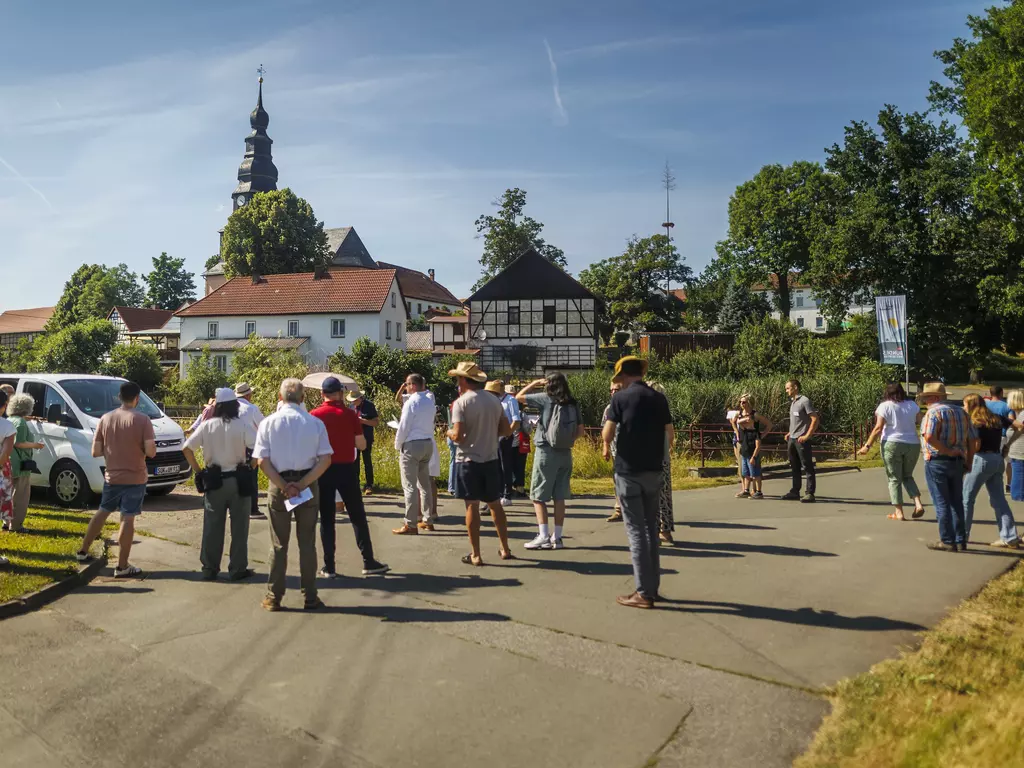 Eine Menschenansammlung steht im Sonnenschein vor einer dörflichen Kulisse und hört einem Redner zu.