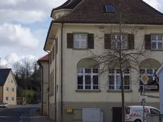 Ein großes leerstehendes altes Gebäude steht rechts am Straßenrand, links kleinere Häuser und eine Kirche. 