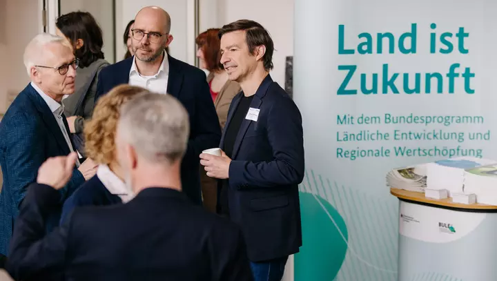 Teilnehmende der Faktor K Konferenz im Gespräch neben einem Roll-up mit der Überschrift "Land ist Zukunft"