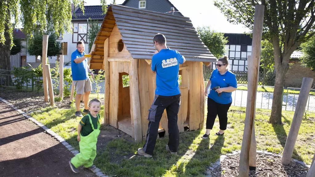 3 Erwachsene und ein Kleinkind werkeln gemeinsam an einer Holzhütte.