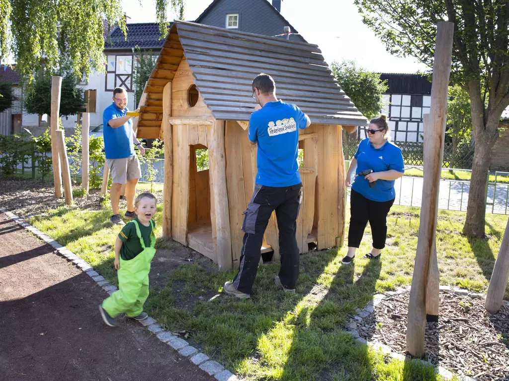 3 Erwachsene und ein Kleinkind werkeln gemeinsam an einer Holzhütte.