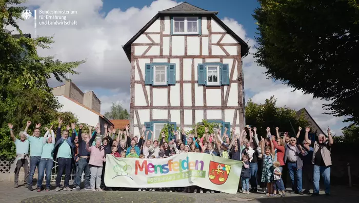 Die Bewohnerschaft des Dorfes Mensfelden steht vor einem Fachwerkhaus und hält gemeinsam ein Banner mit der Aufschrift "Mensfelden - Gemeinsam in die Zukunft" hoch