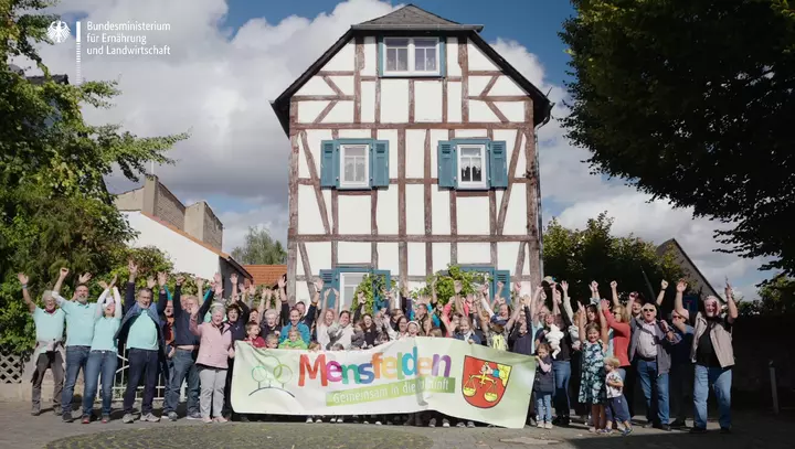 Die Bewohnerschaft des Dorfes Mensfelden steht vor einem Fachwerkhaus und hält gemeinsam ein Banner mit der Aufschrift "Mensfelden - Gemeinsam in die Zukunft" hoch