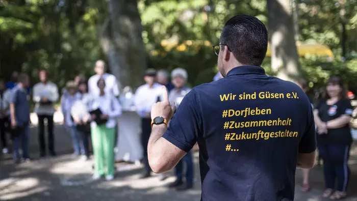 Ein Mann spricht zu im Kreis versammelten Menschen, auf der Rückseite seines Tshirts ist der Aufdruck "Wir sind Güsen e.V."