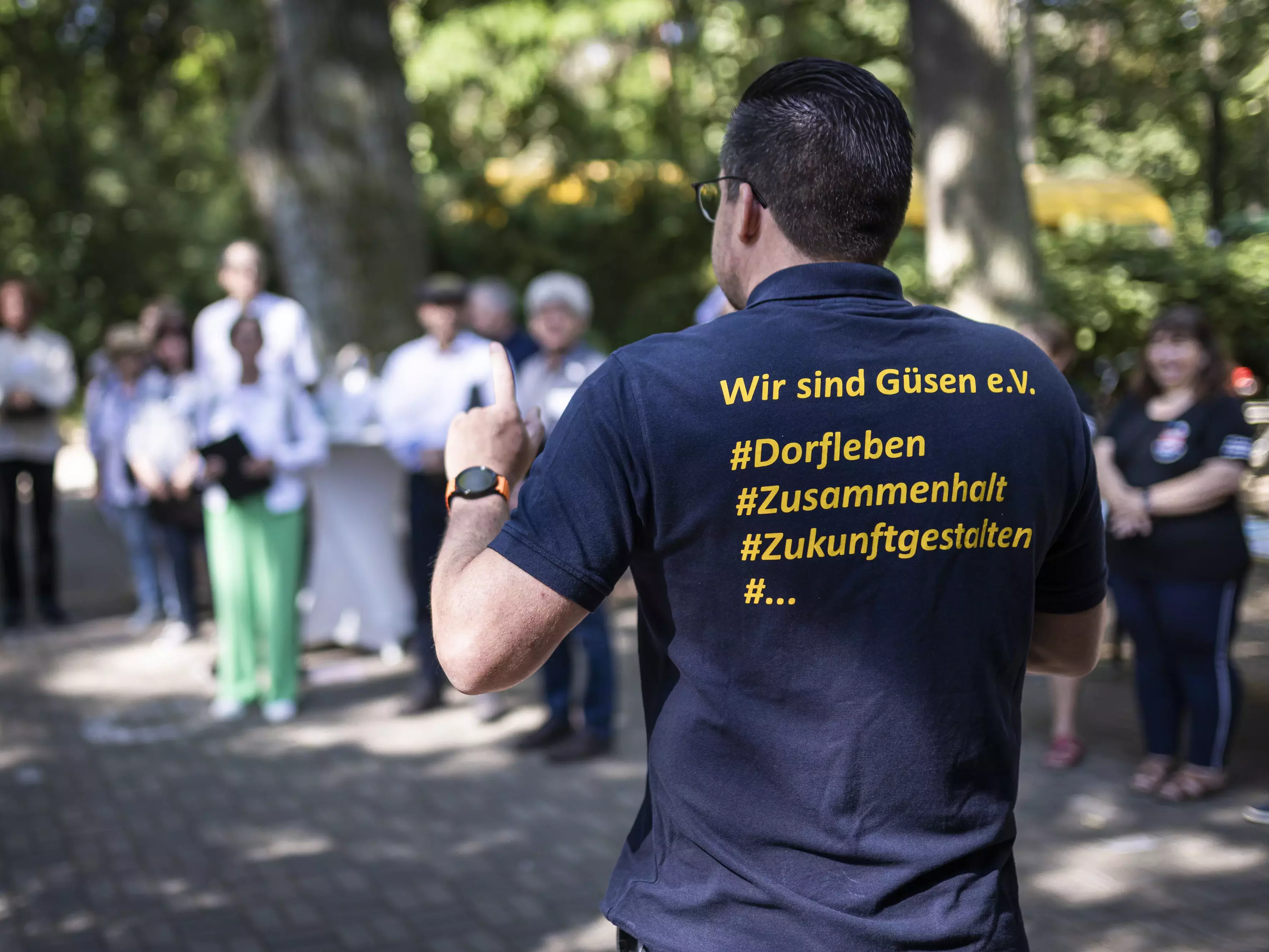 Ein Mann spricht zu im Kreis versammelten Menschen, auf der Rückseite seines Tshirts ist der Aufdruck "Wir sind Güsen e.V."