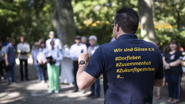 Ein Mann spricht zu im Kreis versammelten Menschen, auf der Rückseite seines Tshirts ist der Aufdruck "Wir sind Güsen e.V."