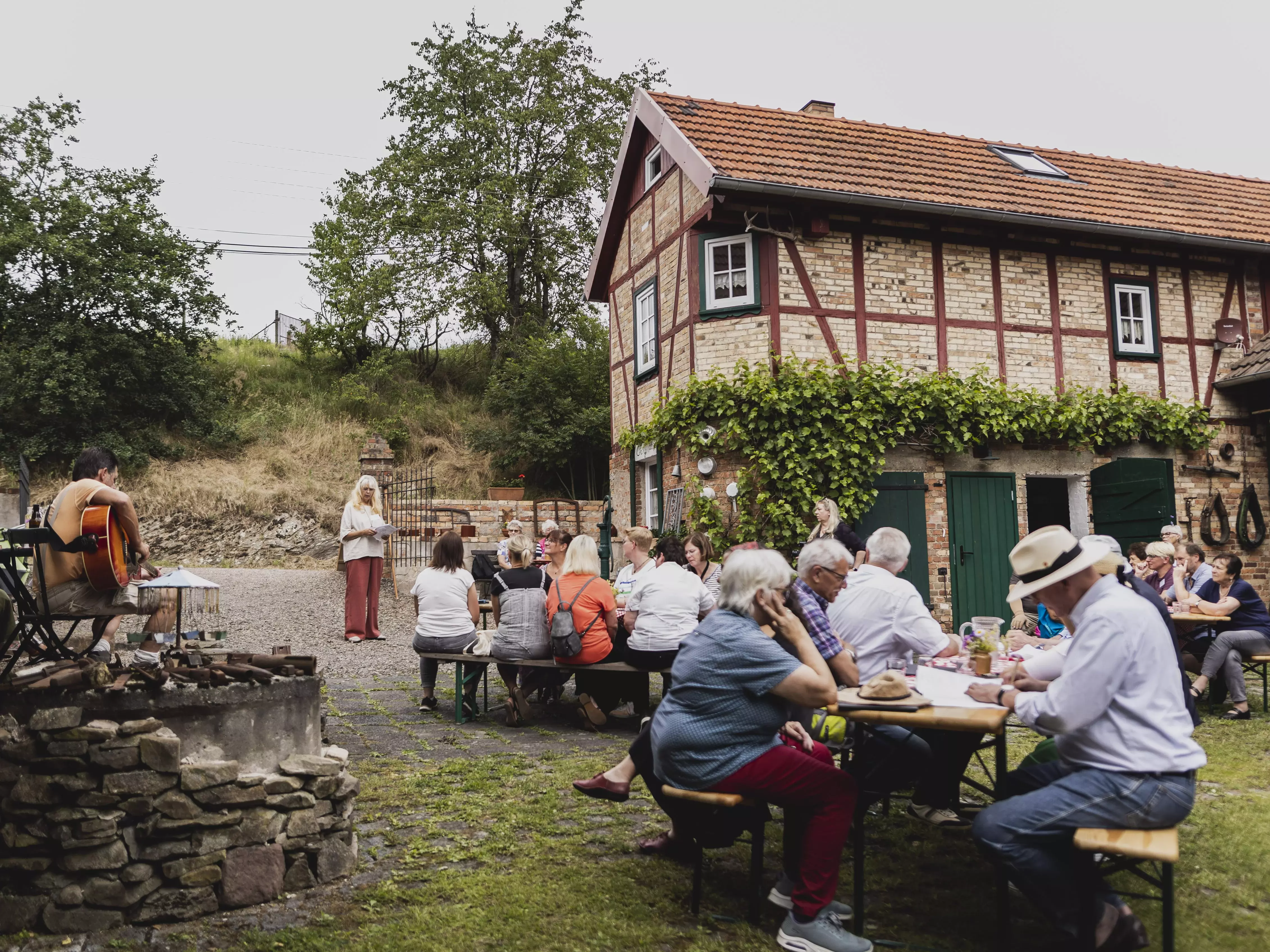 Menschen sitzen auf Bierbänken vor einem Fachwerkhaus.
