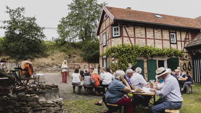 Menschen sitzen auf Bierbänken vor einem Fachwerkhaus.