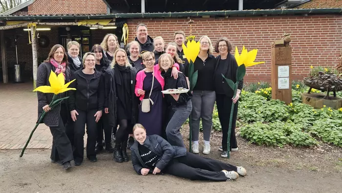 Eine Gruppe von Menschen steht mit großen gebastelten Blumen in der Hand vor einem Haus mit Backsteinfassade. 