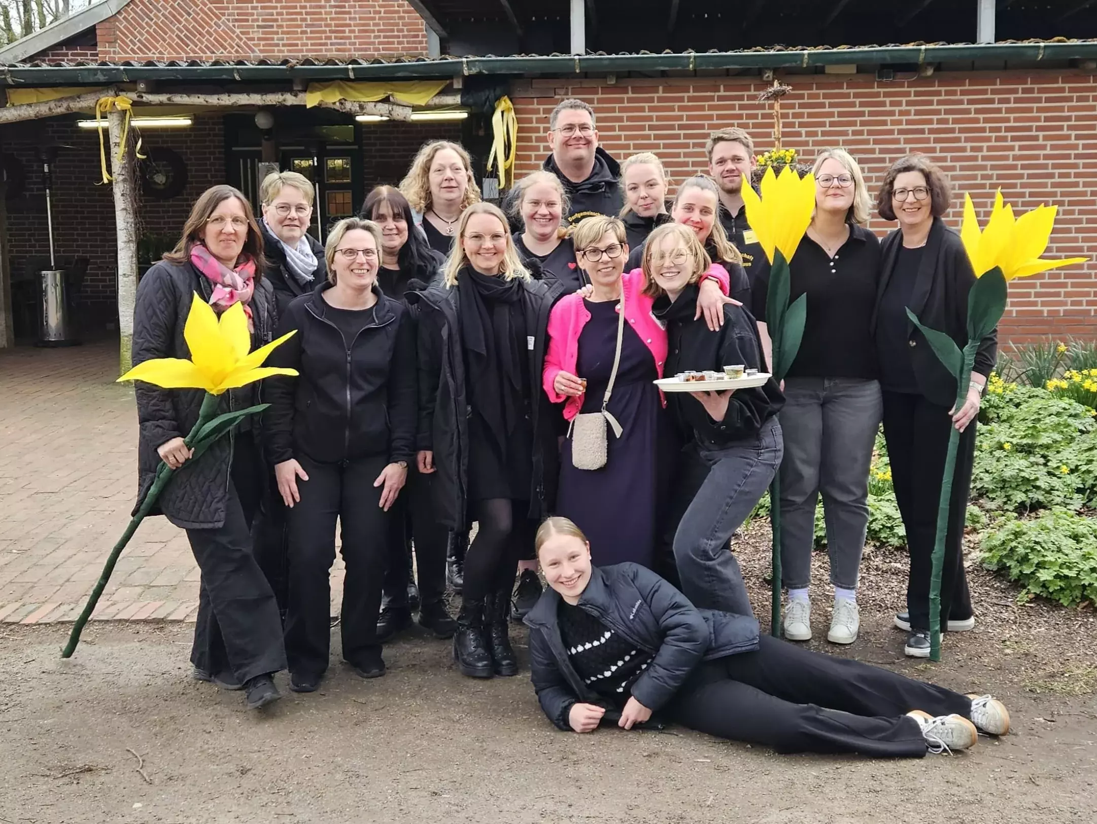 Eine Gruppe von Menschen steht mit großen gebastelten Blumen in der Hand vor einem Haus mit Backsteinfassade. 