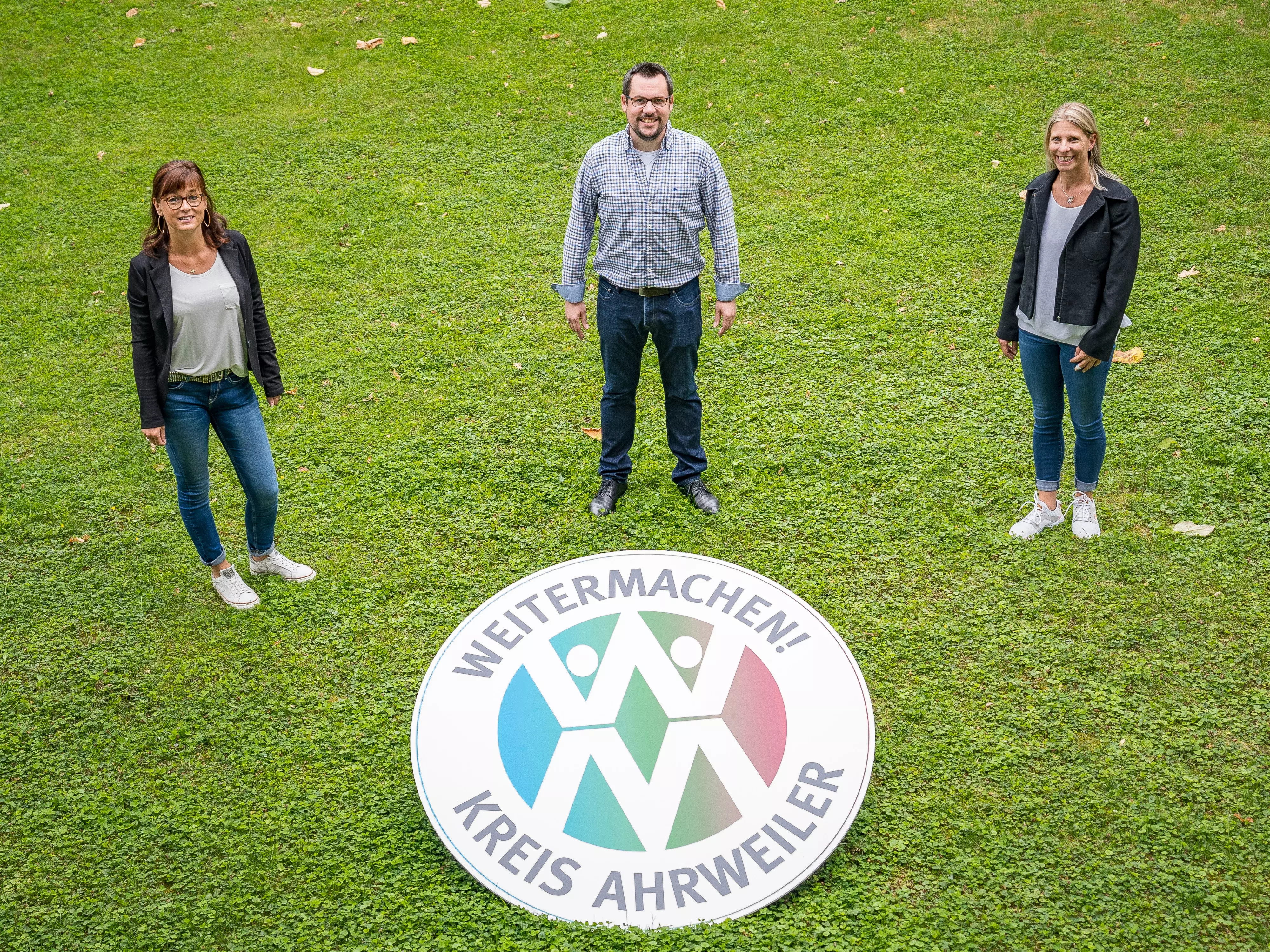 2 Mitarbeiterinnen und 1 Mitarbeiter des Projekts "Weitermachen" stehen mit dem Projektlogo auf einer Wiese