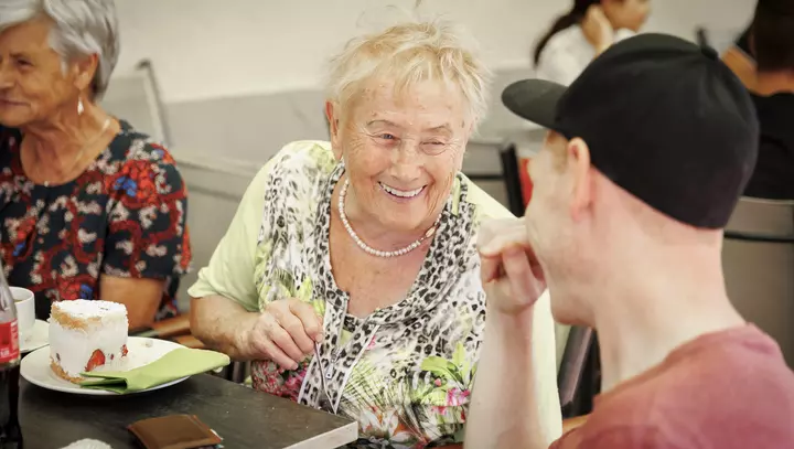 Eine ältere Dame und ein junger Mann mit Kappe sitzen an einem Tisch mit Kuchen