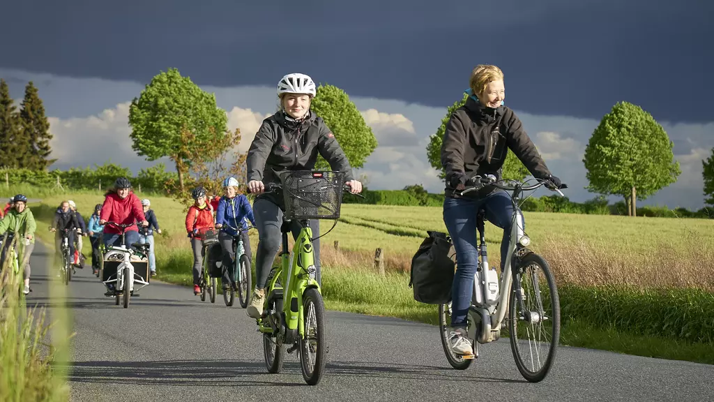 Mehrere Radfahrerinnen sind auf einer Landstraße im Sonnenschein unterwegs.