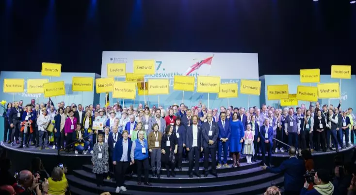 Teilnehmende des Dorfwettbewerbs stehen mit ihren Dorfschildern zusammen mit Bundesminister Özdemir  bei der Abschlussfeier auf der Bühne  
