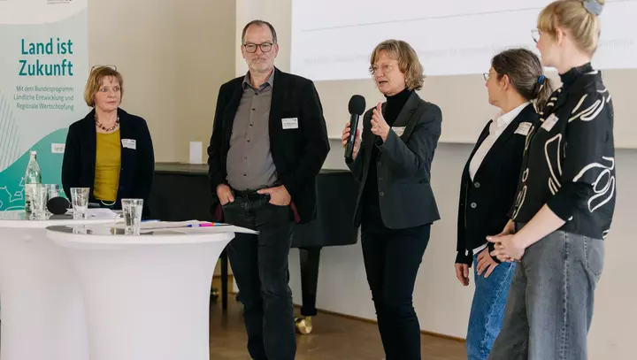 5 Personen stehen hinter einem Stehtisch, eine Frau hält ein Mikrofon in der Hand