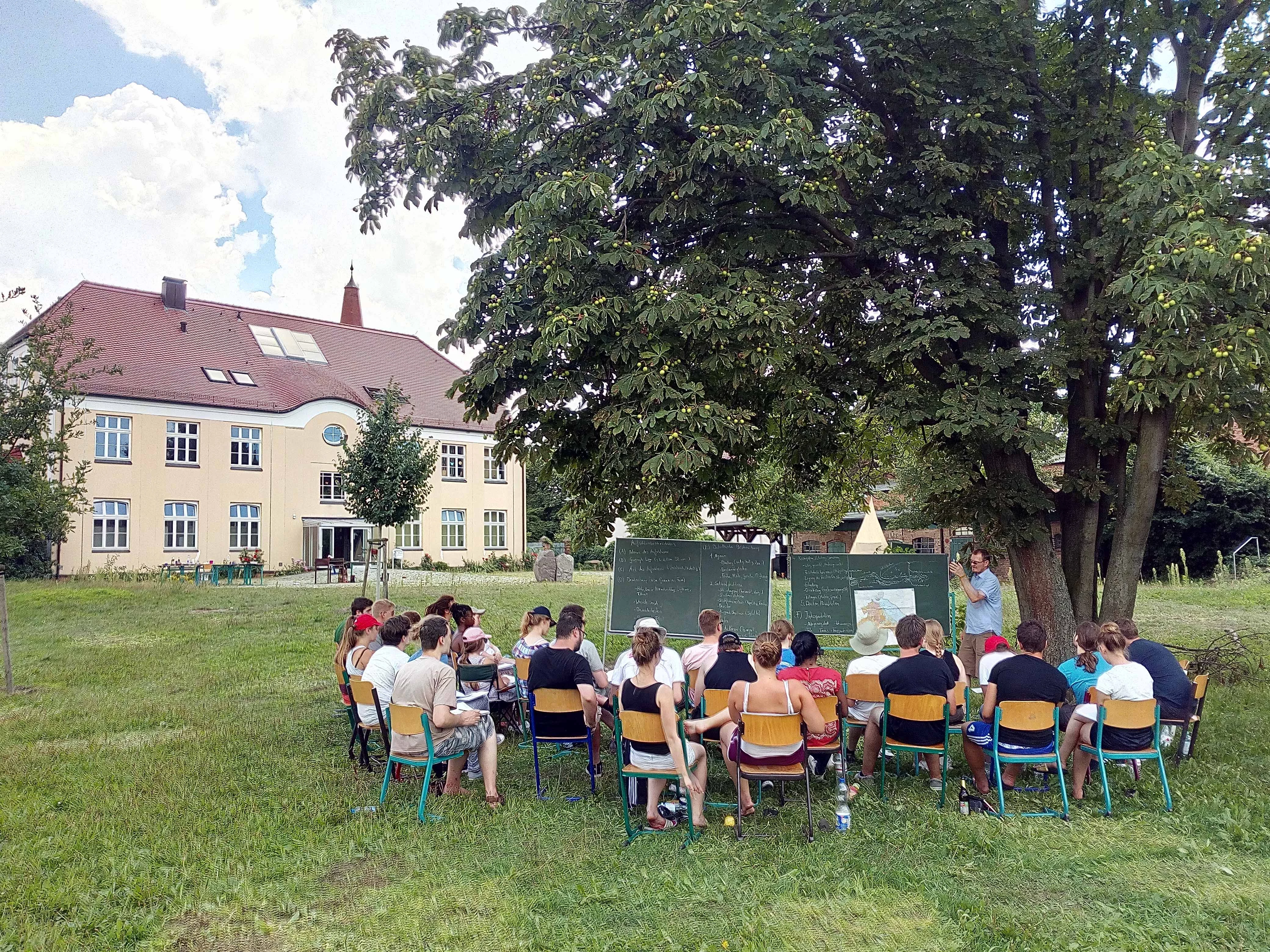 Menschen sitzen auf Stühlen vor einer Tafel auf einer Wiese