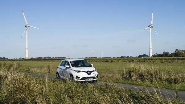 Ein Auto fährt über eine Landstraße, im Hintergrund sind zwei Windräder zu sehen.