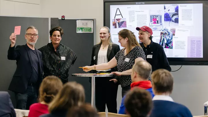 5 Personen stehen um einen Stehtisch. Eine Person hält eine Spielkarte in die Höhe.