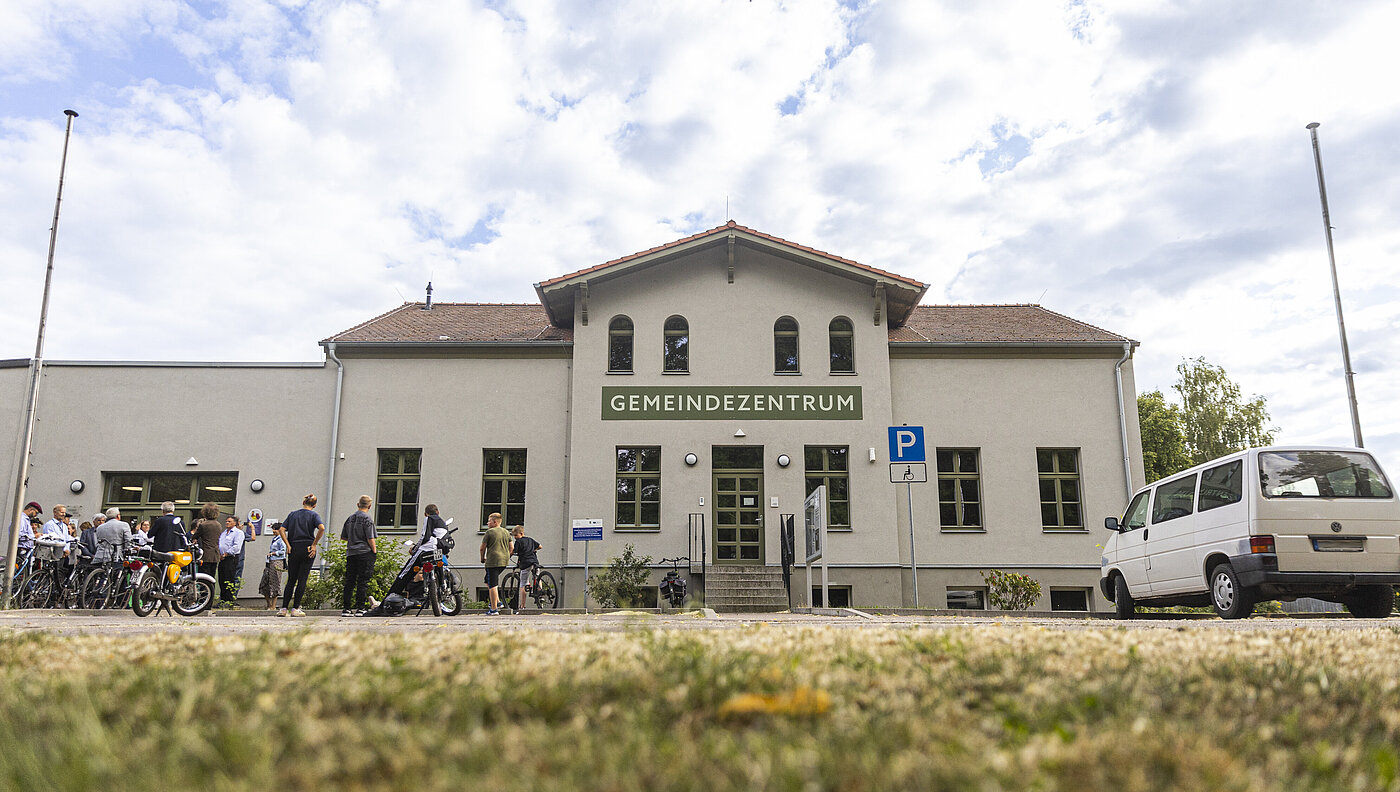 Frontansicht eines Gemeindezentrums, links davor befinden sich Menschen mit Fahrrädern und Rollern
