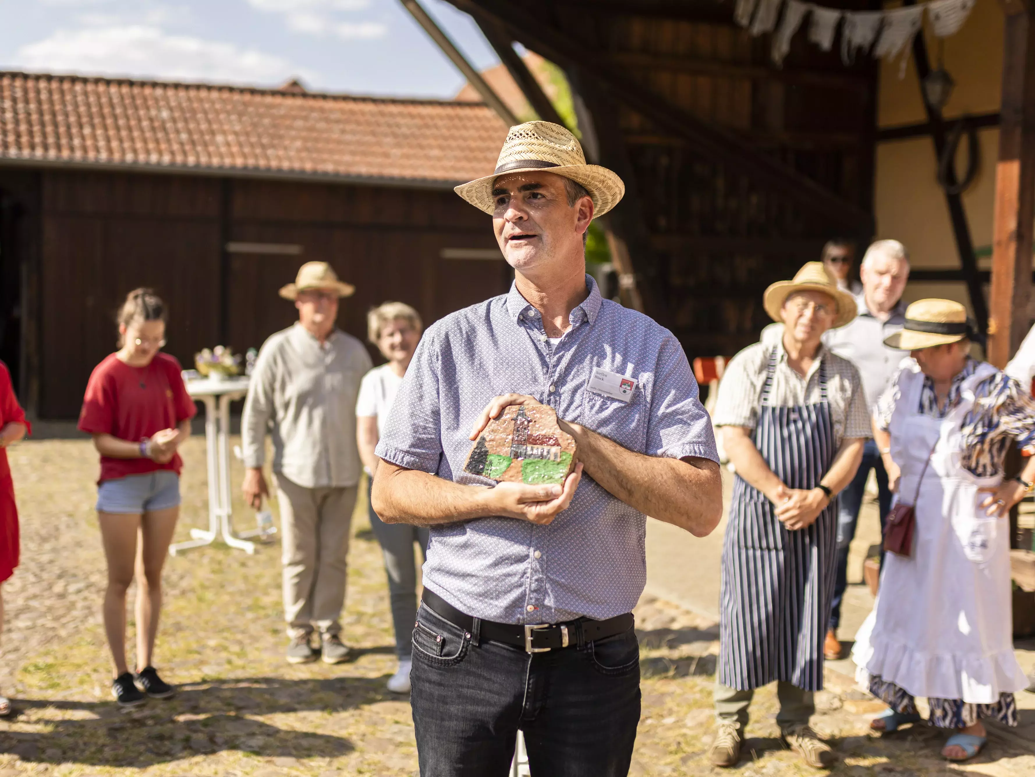 Ein Mann steht vor einem Halbkreis aus Menschen und hält eine bemalte Tonplatte in der Hand. 