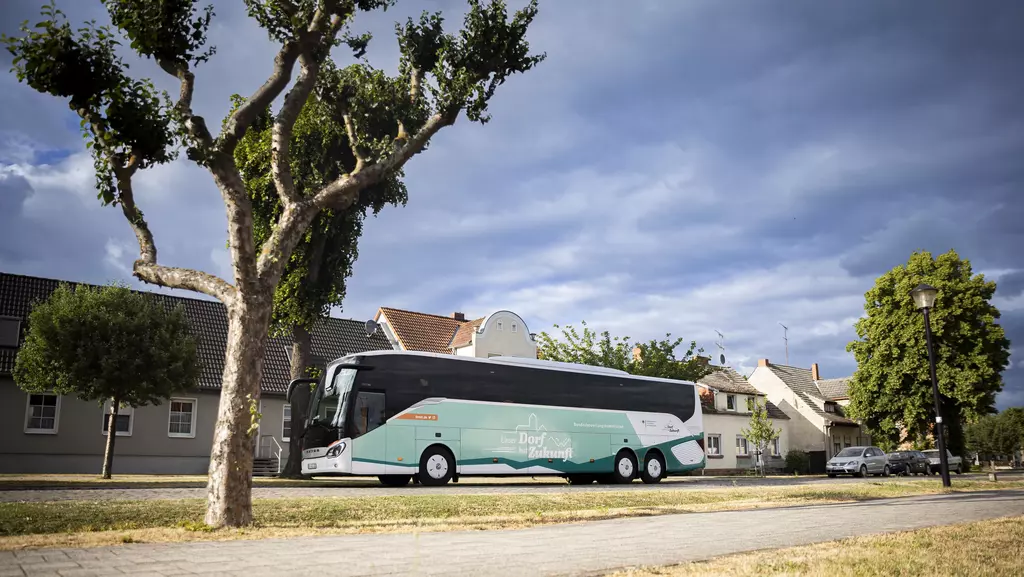 Ein Bus mit Logo und Schriftzug des Bundeswettbewerbs "Unser Dorf hat Zukunft" auf einer Straße mit Bäumen