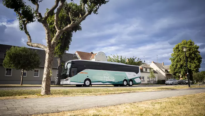 Ein Bus mit Logo und Schriftzug des Bundeswettbewerbs "Unser Dorf hat Zukunft" auf einer Straße mit Bäumen